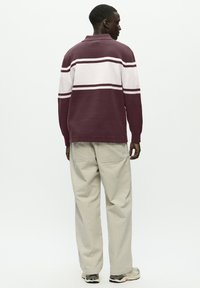 Person iført en maroon og hvid stribet langærmet skjorte, beige bukser og sneakers, stående med ryggen til på en ensfarvet baggrund.