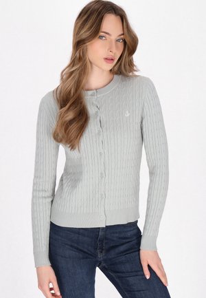 Cardigan a coste grigio chiaro con scollo rotondo, chiusura a bottoni e piccolo emblemma di ancora. Indossato con jeans blu scuro.