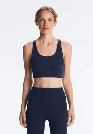 Ženska v temno modrem športnem modrčku in gamašah z visokim pasom stoji z rokami sproščeno ob telesu na svetlo sivi podlagi.