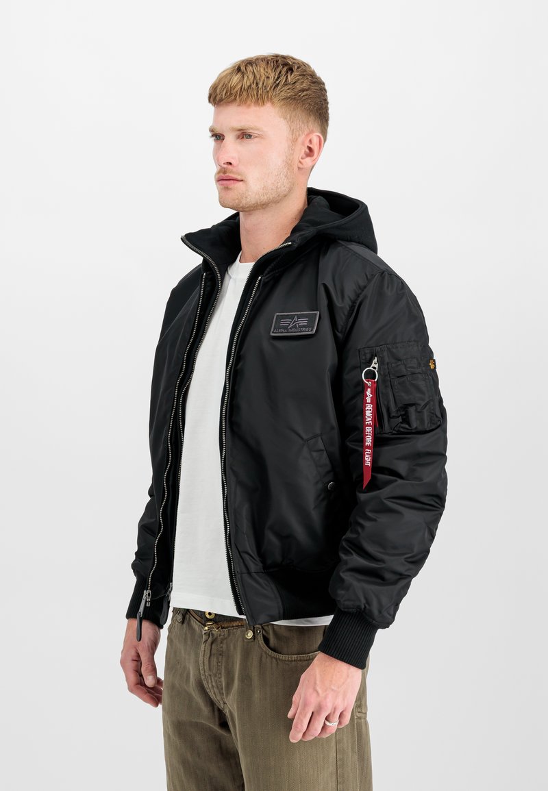Sort bomberjakke med aftagelig hætte, lynlås foran, ribbede manchetter og patchdetalje på brystet. Inkluderer sidelommer og dekorativ flap.