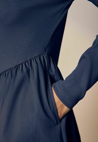 Navyblauwe jurk met geribbelde lijfje, lange mouwen en geplooide taille, die soepel overgaat in een vloeiende rok met zijzak.