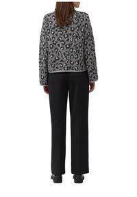 Vrouw in een grijs luipaardprint trui, zwarte rechte broek en zwarte schoenen, staande met de rug naar de camera tegen een witte achtergrond.