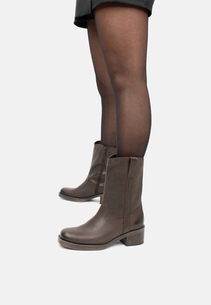 Botines de cuero marrón con punta cuadrada, cremallera lateral y tacón apilado, mostrados con medias negras transparentes.