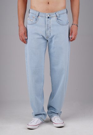 Mann in hellblauen, weit geschnittenen Jeans und weißen Sneakers, der vor einem schlichten grauen Hintergrund steht, oberkörperfrei mit einem Armband am rechten Handgelenk.