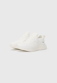 Zapatillas deportivas blancas con parte superior de malla, puntera redonda, cordones planos y suela de goma texturizada con una ligera elevación.