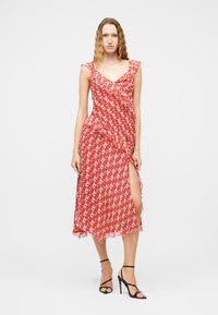 Γυναίκα που φοράει midi φόρεμα χωρίς μανίκια, κόκκινο και ροζ με σχέδια, βολάν και σχίσιμο μέχρι το μηρό, συνδυασμένο με μαύρα τακούνια με λουράκια.