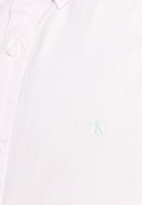 Camicia rosa chiaro con colletto, realizzata in tessuto morbido, con un discreto logo "cK" verde ricamato sul petto. Design minimalista, senza motivi.