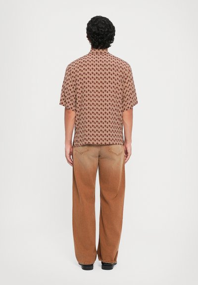 Kurzarmhemd mit einem braunen geometrischen Muster, kombiniert mit weit geschnittenen, braunen Hosen, beide aus strukturiertem Stoff. Schwarze Schuhe.