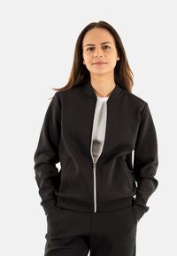 Giacca bomber nera con zip, dal tessuto liscio, maniche a costine e colletto tondo. Indossata sopra una maglietta bianca aderente, è elegante e funzionale.