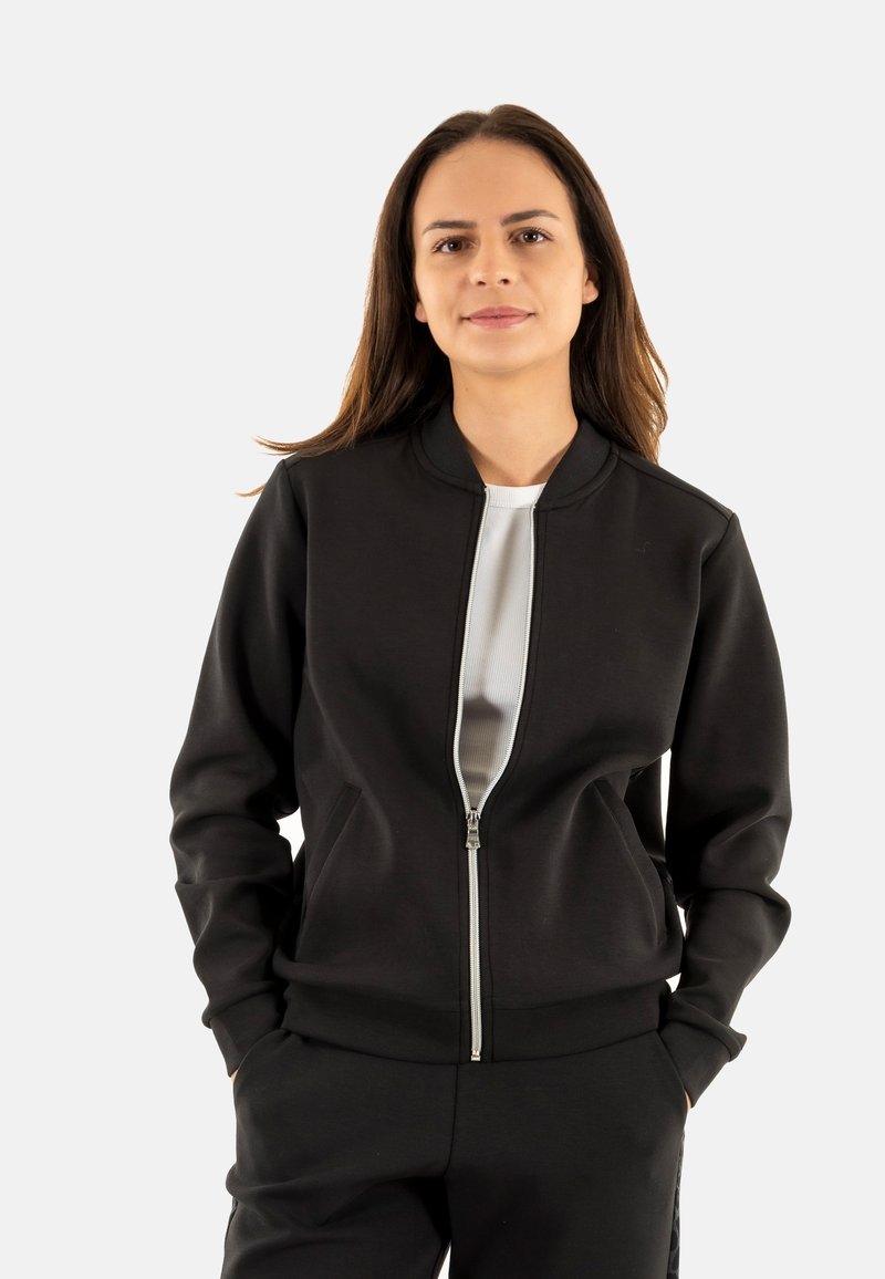 Giacca bomber nera con zip, dal tessuto liscio, maniche a costine e colletto tondo. Indossata sopra una maglietta bianca aderente, è elegante e funzionale.