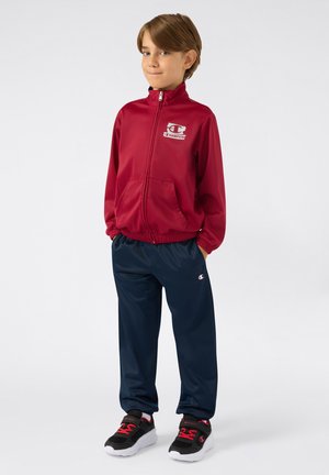 Chaqueta roja con cremallera y logo, pantalones de cintura elástica azul marino, y zapatillas negras con detalles rojos. Tejido suave, corte atlético, diseño práctico.