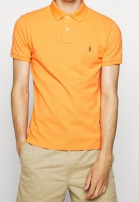 Orange polo shirt i bomull, med krage, tvåknappssläpp och en liten grön logotyp på vänster bröst. Enfärgad textur.
