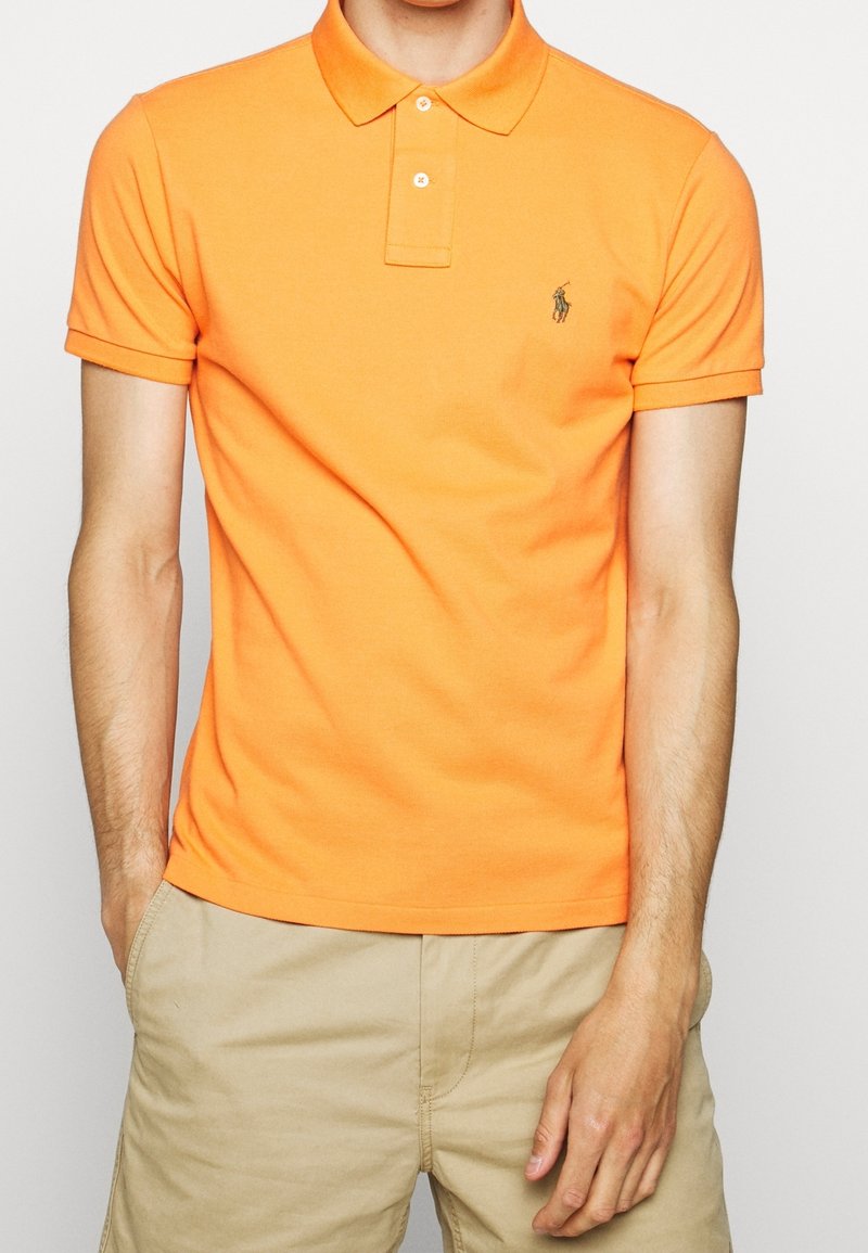 Orange polo shirt i bomull, med krage, tvåknappssläpp och en liten grön logotyp på vänster bröst. Enfärgad textur.