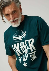 Camiseta de algodón de color teal con un gran gráfico blanco de un sombrero de marinero con un ancla y texto divertido "Bajo el Mar". Manga corta, corte relajado.