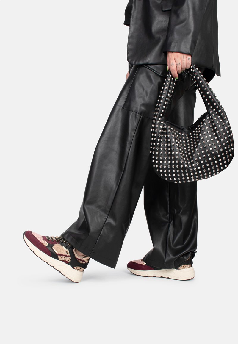 Un pantalon oversize en simili cuir noir associé à des sneakers à motif dans des tons bordeaux, noir et beige. Un sac à main noir clouté vient parfaire le look.