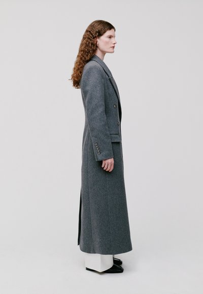 Cappotto lungo grigio in lana con una struttura ben definita, chiusura a un solo bottone e uno spacco laterale, abbinato a pantaloni bianchi e scarpe nere.