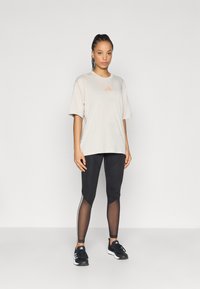 Φαρδιά φανέλα σε ανοιχτό μπεζ χρώμα με πορτοκαλί λογότυπο, συνδυασμένη με μαύρα κολάν με διάτρητες λεπτομέρειες και λευκές ρίγες στο πλάι, φορεμένη με μαύρα αθλητικά παπούτσια.