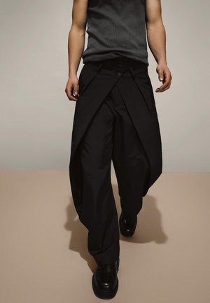 Férfi fekete aszimmetrikus, széles szárú nadrágot, fekete cipőt és sötét ujjatlan felsőt visel, bézs padlón áll, semleges fal előtt.