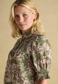 Blond naine kergete pintslitega kannab roheline ja roosa mustriga pluusi, millel on puhvlivarrukad, seistes beeži taustal.