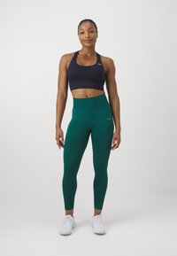 Black sports bra and dark green high-waisted leggings, made from stretchy fabric, paired with white athletic shoes. Simple logo detail visible.