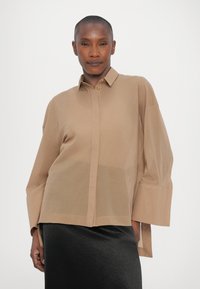 Light brown button-up shirt with long sleeves, a relaxed fit, and a semi-sheer material, paired with a black textured skirt.