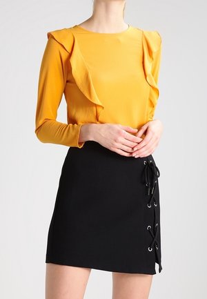 Gul langærmet top med flæseaccenter, parret med sort mininederdel med snøredetalje i siden. Glat stoftekstur.