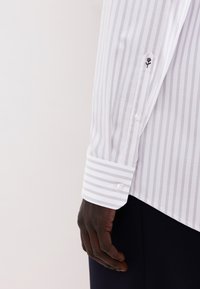 Camicia a maniche lunghe bianca a righe con un piccolo ricamo di rosa nera sul polsino, caratterizzata da una classica chiusura con bottoni e tessuto strutturato.