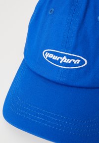 Blå bomullskaps med en strukturert design, buet skygge og brodert "yourturn"-logo i hvitt på fronten. Teksturert stoff med synlige sømmer.