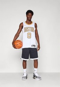 Noor mees Bostoni korvpallimeeskonna särgis ja lühikestes pükstes, hoides korvpalli, seisab neutraalses heledas taustas.