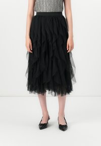 Kvinne iført et lagvis svart tyll midi-skjørt, grå kabelstrikket ermeløs topp og svarte spisse slingback hæler, som står mot hvit bakgrunn.