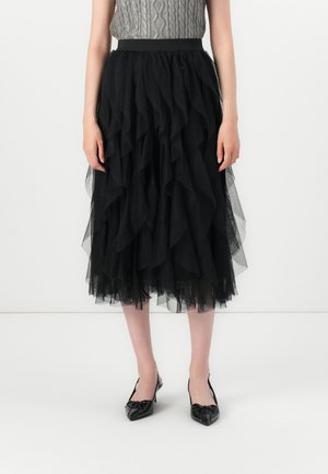 Woman wearing a layered black tulle midi skirt, gray cable-knit sleeveless top, and black pointed-toe slingback heels, standing against white.