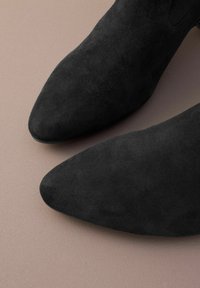 Bottines en daim noir à bout pointu. Présentent une texture lisse et un style minimaliste, affichées sur un fond neutre.
