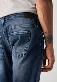 Blugi de denim de culoare albastru închis, cu o textură uzată, având două buzunare pe spate și o eticheta din piele neagră la talie.