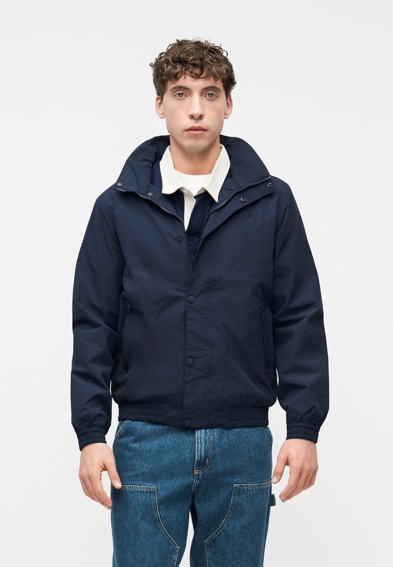 Νέος άνδρας που φοράει μια ναυτική μπλε ζακέτα με κουκούλα πάνω από λευκό πουκάμισο με γιακά και μπλε τζιν, στέκεται μπροστά σε απλό φωτεινό φόντο.