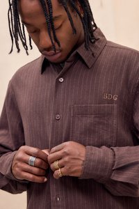 Camicia marrone con bottoni, decorata con strisce verticali bianche, presenta una tasca sul petto, colletto e dettaglio ricamato "BDG". Mani adornate con anelli.