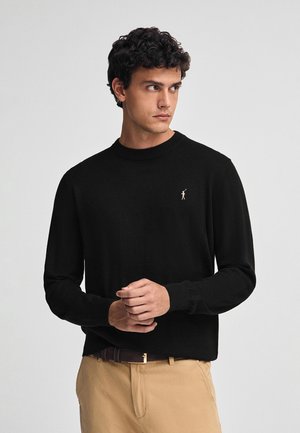 Pull en tricot noir avec un col rond et des manches longues. Présente un petit logo brodé discret sur la poitrine.