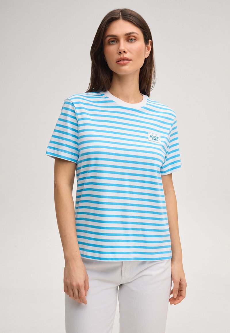 Femme portant une chemise à manches courtes rayée blanc et bleu clair avec le logo « JOOP! JEANS » sur la poitrine, associée à un pantalon blanc.