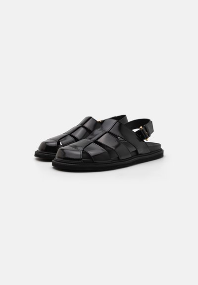 Black sandals made of glossy leather featuring a woven design, open-toe, and an adjustable strap with gold hardware. Flat sole.