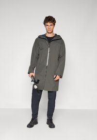 Parka gris resistente al agua con cierre de cremallera, capucha ajustable y mangas largas, combinada con pantalones oscuros y zapatos negros. Sostiene un par de bastones de senderismo.