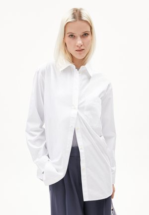 Chemise blanche à manches longues avec un bouton et une poche, dotée d'un col classique et d'une texture lisse, associée à un pantalon foncé.
