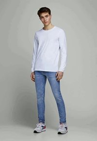Vit långärmad skjorta, ljusblå skinny jeans och vita sneakers med röda och blå accenter. Modellen står mot en enkel bakgrund.