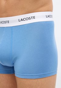 Κοντινό πλάνο ενός ατόμου που φορά ελαφρώς μπλε μπόξερ Lacoste με λευκή μέση που διαθέτει το λογότυπο Lacoste σε μαύρο χρώμα.