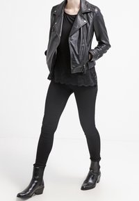 Person som bär en svart läderjacka, en svart topp med spetsdetaljer, svarta tighta byxor och svarta ankelstövlar med klack, står med händerna i fickorna.