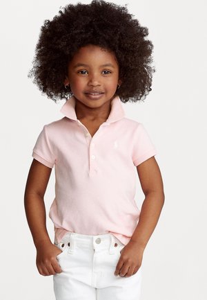 Camisa polo rosa con cuello, mangas cortas y un pequeño logo. Combinada con pantalones blancos. Textura suave y diseño clásico.