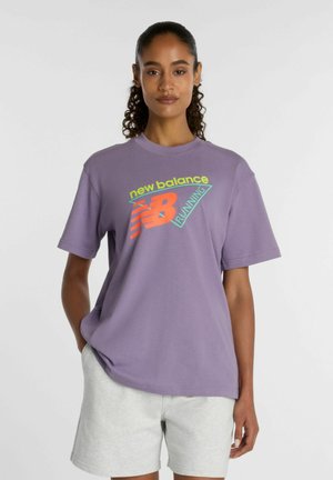 Kvinde iført en lilla New Balance løbe-t-shirt og lysegrå shorts, stående med den ene hånd i lommen mod en ensfarvet baggrund.