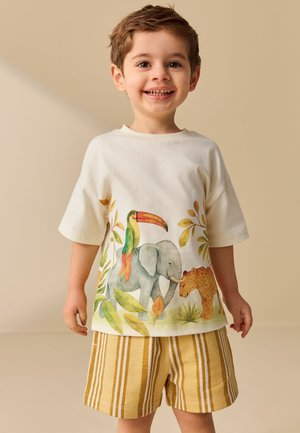 Junger lächelnder Junge, trägt ein weißes T-Shirt mit bunten Dschungeltieren und senfgelbe gestreifte Shorts, steht vor einem beigen Hintergrund.
