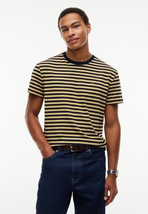 Ung mand iført en marineblå og gulstribet T-shirt med mørkeblå jeans, en hånd i lommen, stående mod en ensfarvet hvid baggrund.