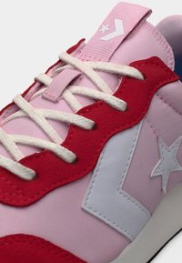 Ροζ αθλητικό παπούτσι με λευκά κορδόνια, κόκκινες επενδύσεις σουέτ και λευκό αστέρι και λογότυπο σήματα βέλους στο πλάι και στη γλώσσα.