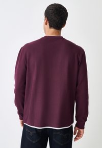 Langärmeliges Pullover in Kastanienbraun aus strukturiertem Stoff, mit Rundhalsausschnitt, legerer Passform und geschichtetem Saum, kombiniert mit dunklen Jeans.