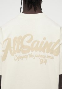 Μπεζ βαμβακερό t-shirt με μια μεγάλη, μπεζ γραφική επιγραφή στην πλάτη που γράφει "AllSaints" με τη φράση "Απολαμβάνοντας το ταξίδι από το '94."